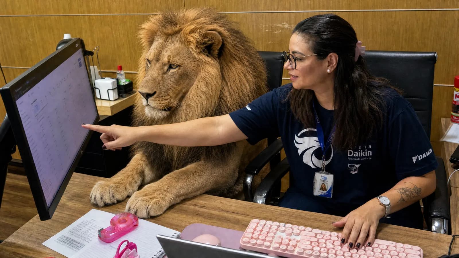 Colaboradora mostra a planilha no monitor enquanto um leão, representando o agente de IA, observa com atenção